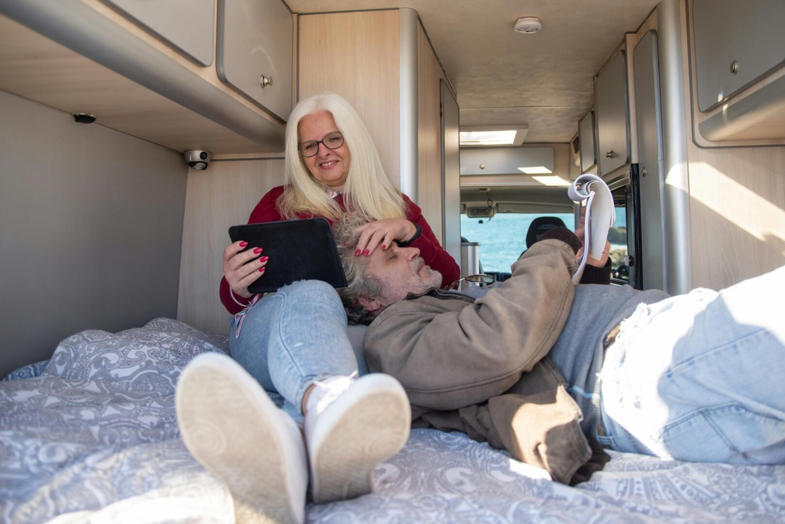 A senior couple enjoys a relaxing day inside their camper van, reading and using a tablet.